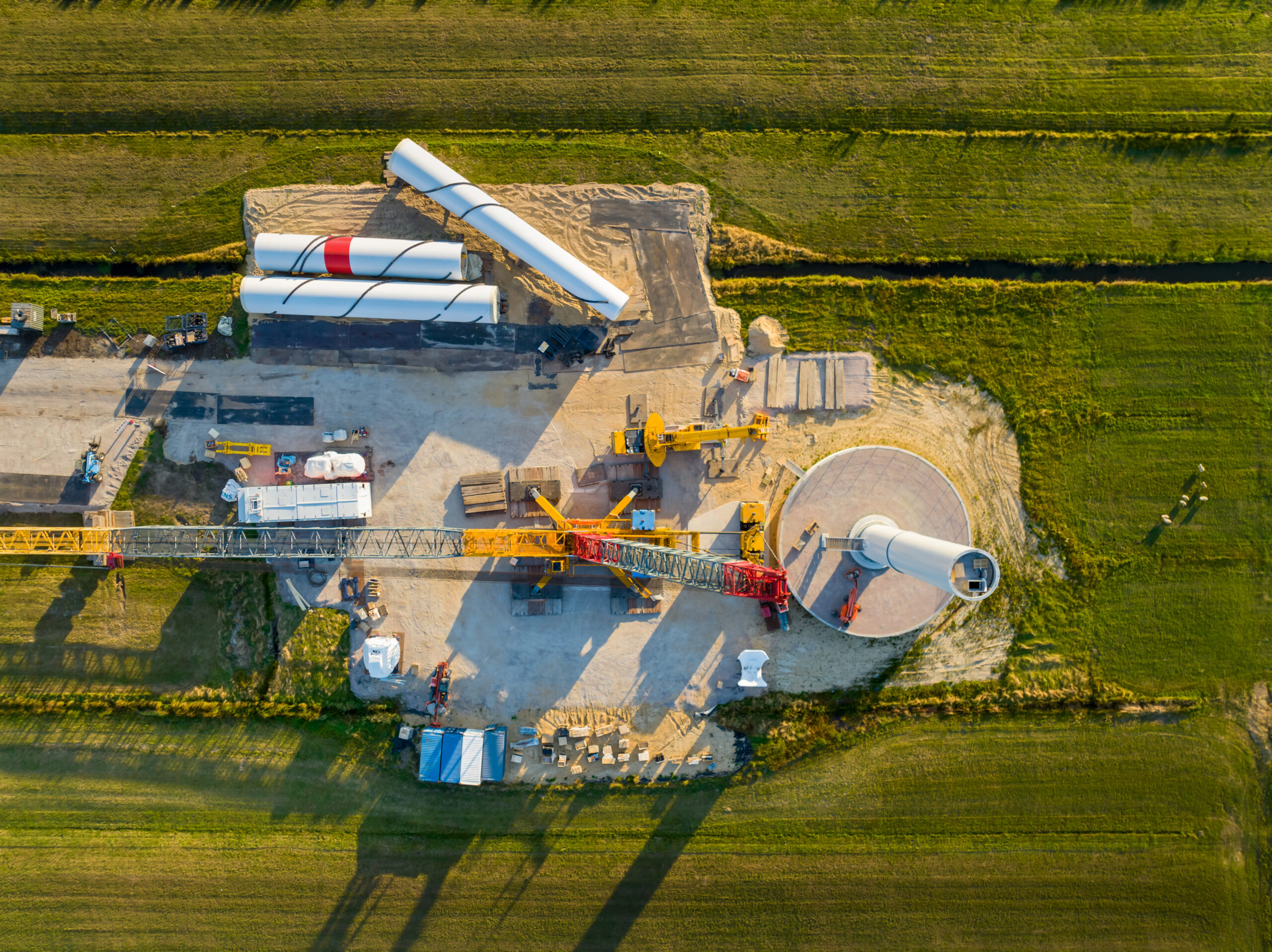 Aerial view of a wind turbine construction site showing a large crane, turbine tower base, and three blades staged on the ground awaiting installation, surrounded by green agricultural fields.