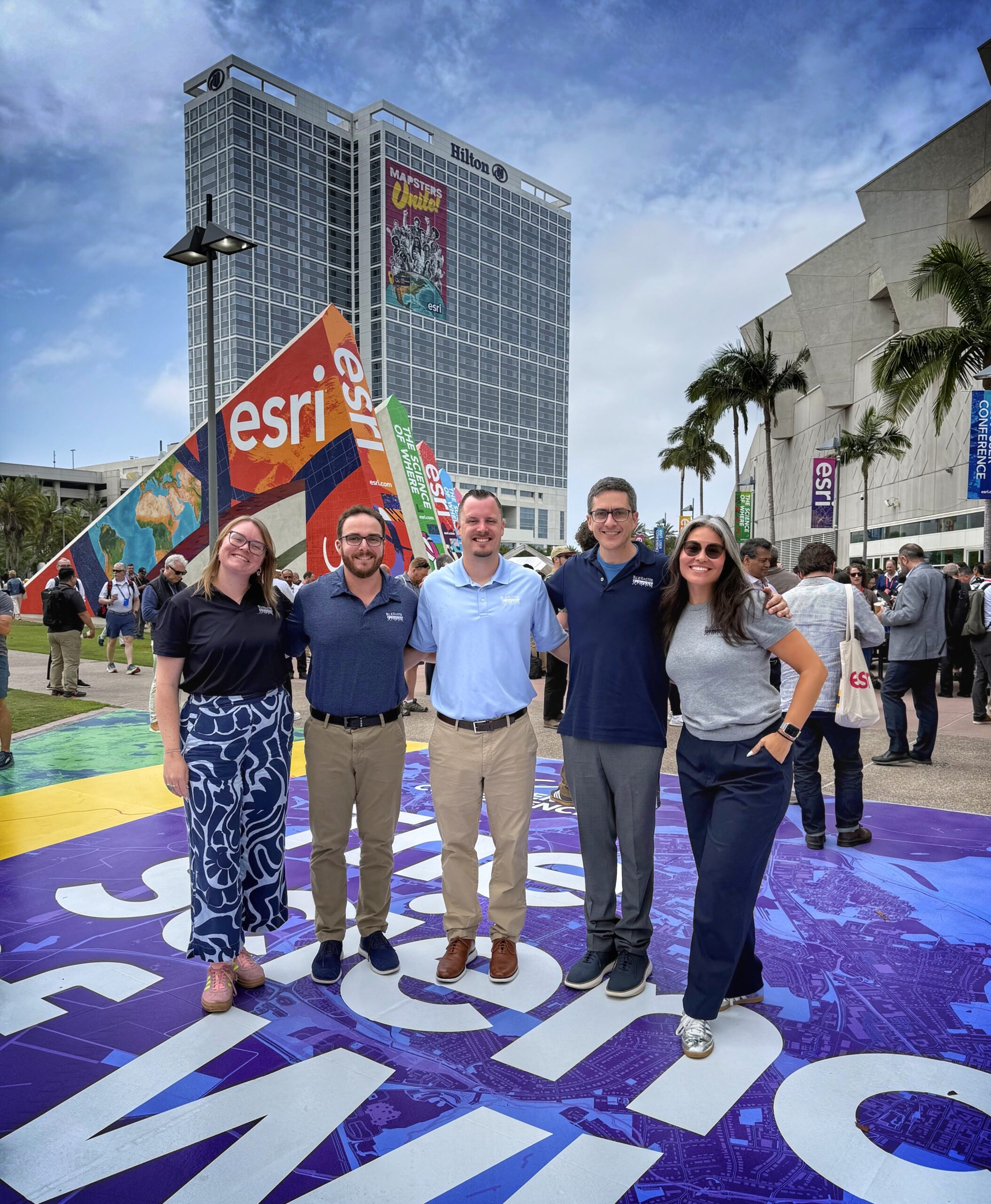 Five members of the Blue Raster team posing in front of the Esri signage and Hilton hotel outside the San Diego Convention Center at the 2026 Esri User Conference, with the Mapsters Unite banner visible in the background
