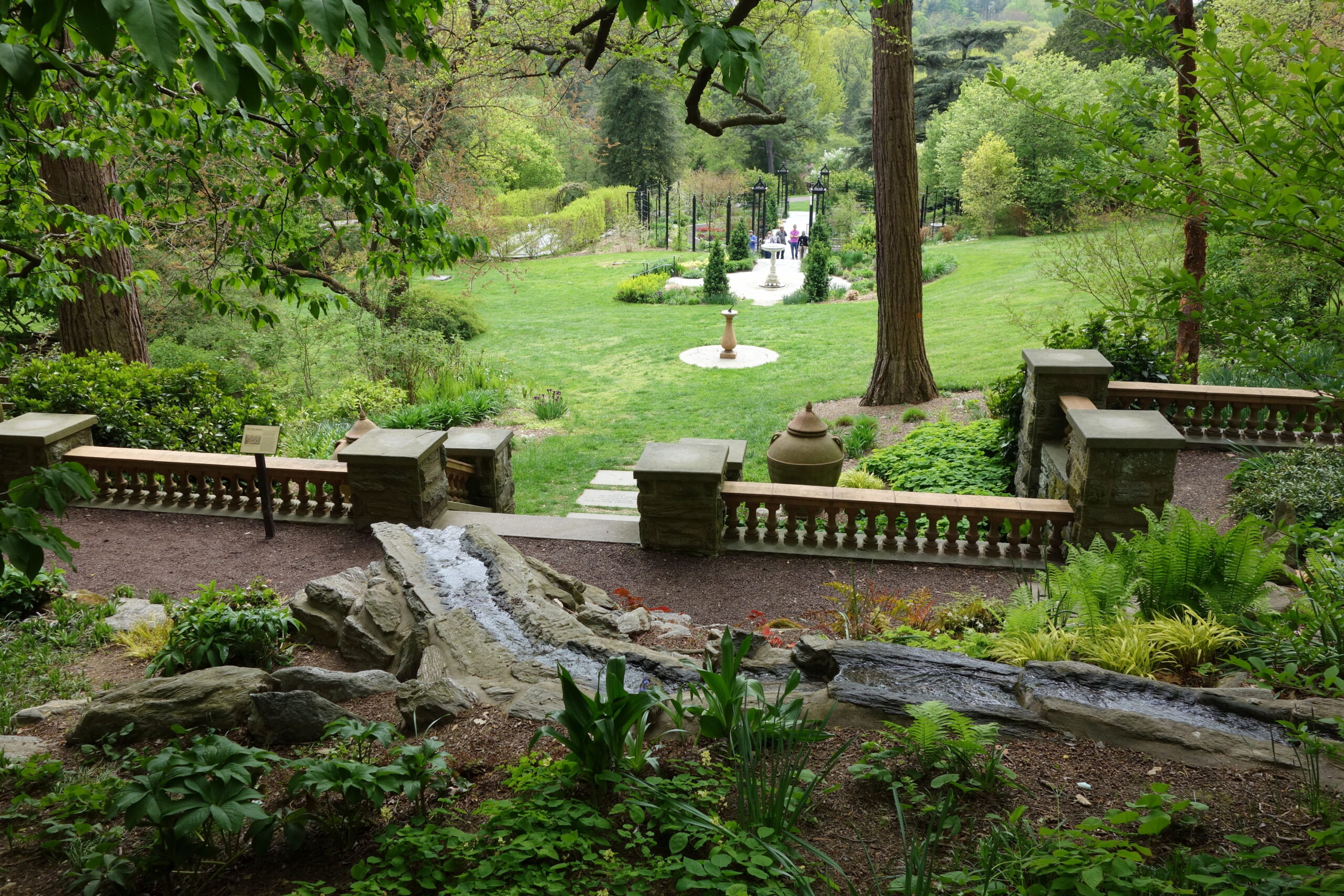 View from the upper terrace at Morris Arboretum & Gardens looking down stone balustrade steps toward a sundial fountain, open lawn, and tree-lined garden path in Philadelphia's Chestnut Hill neighborhood