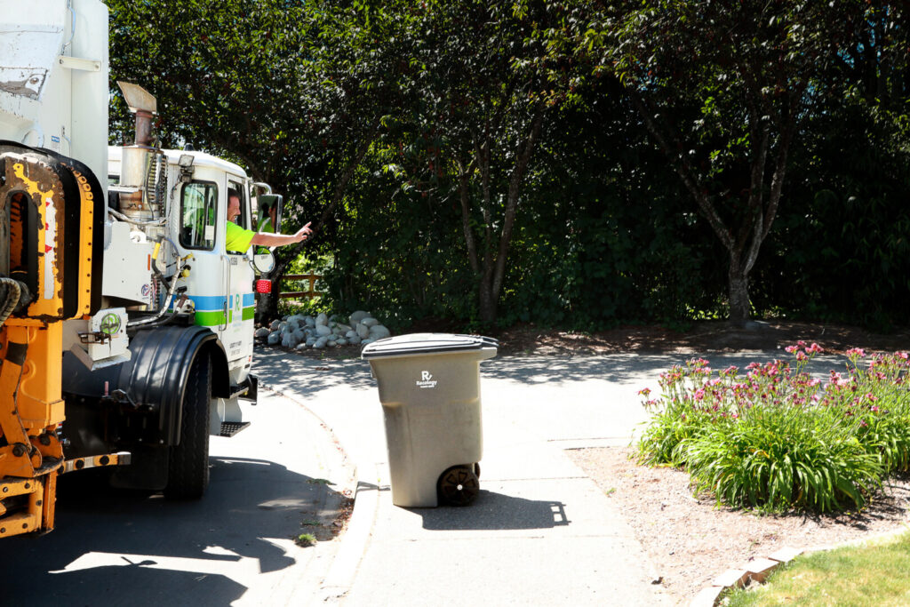 Photo of a Recology garbage truck driver, wearing a high-visibility green shirt, leaning out of the cab window and gesturing with his hand while operating the truck's automated arm on a residential street. A gray Recology waste bin sits on the sidewalk in front of the truck, positioned to be picked up by the mechanical arm. The background features lush green trees, a small rock garden, and a flower bed with pink blooms on the right side of the driveway.