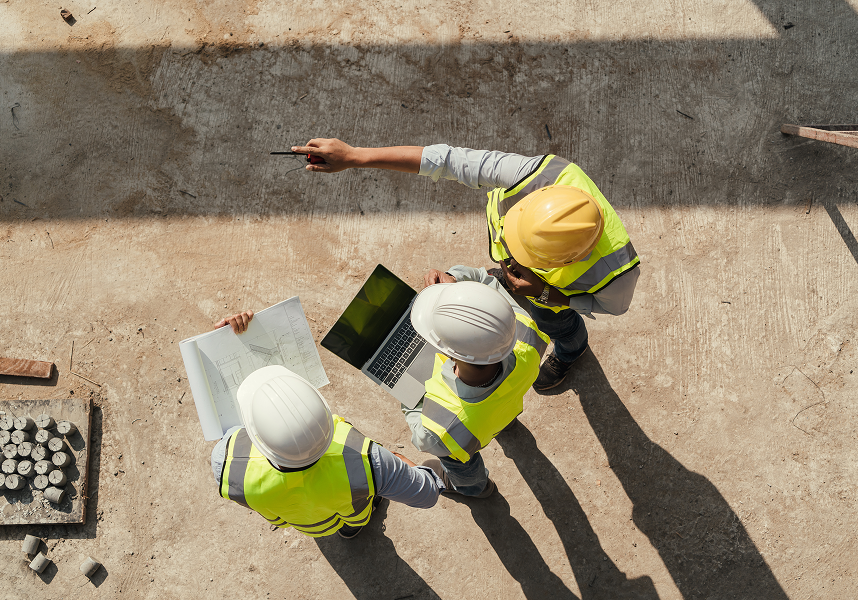 Overhead view of three construction workers in hard hats and high-visibility vests reviewing blueprints and a laptop on a construction site.