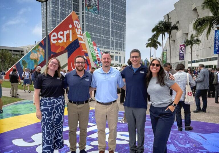 Five members of the Blue Raster team posing in front of the Esri signage and Hilton hotel outside the San Diego Convention Center at the 2026 Esri User Conference, with the "Mapsters Unite" banner visible in the background