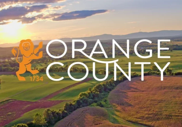 Orange County, Virginia official branding image featuring the county logo with a lion mascot and the year 1734, overlaid on an aerial sunset view of rolling farmland, green fields, and the Blue Ridge Mountains in the background.