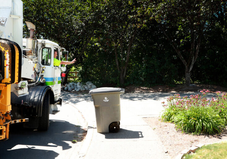 Photo of a Recology garbage truck driver, wearing a high-visibility green shirt, leaning out of the cab window and gesturing with his hand while operating the truck's automated arm on a residential street. A gray Recology waste bin sits on the sidewalk in front of the truck, positioned to be picked up by the mechanical arm. The background features lush green trees, a small rock garden, and a flower bed with pink blooms on the right side of the driveway.